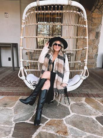 A reviewer relaxing in an egg chair, sporting a black hat, sunglasses, a beige scarf, a beige top, and black knee-high boots, striking a chic pose for the camera.