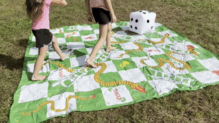 Two children enjoy playing on a large outdoor snakes and ladders mat, with an oversized inflatable die placed on the grass.