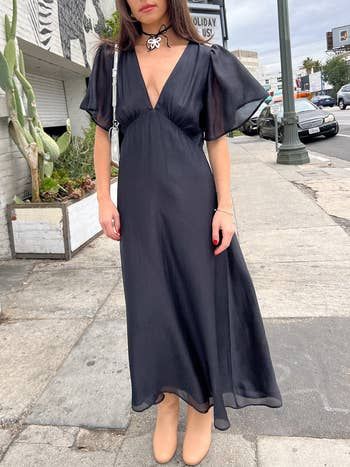 A person in a flowing black dress paired with a butterfly choker necklace, standing gracefully on a city sidewalk.