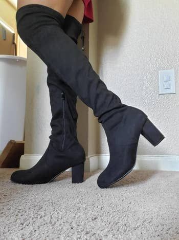 A person stands indoors on a carpet, wearing thigh-high black boots with heels.
