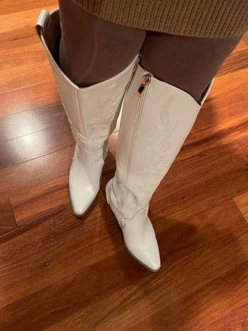 A close-up shot of a reviewer wearing white, knee-high boots with intricate stitching, paired with brown leggings and a brown skirt, standing on a wooden floor.