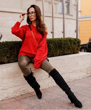 A model sits on a bench outside, wearing glasses, a red sweater, olive pants, black knee-high boots, and carrying a black purse with a gold chain strap