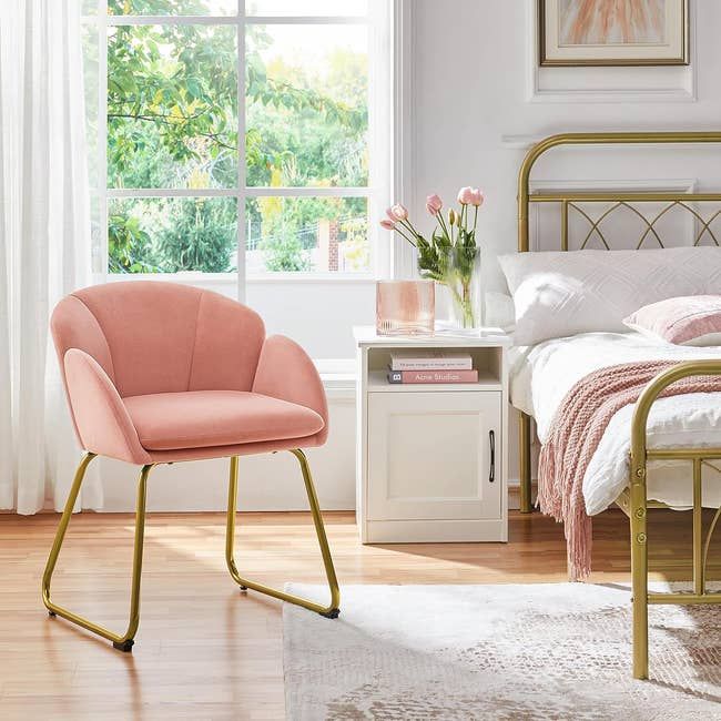 A beautiful pink armchair with gold legs next to a bed with a white nightstand and pink flowers.