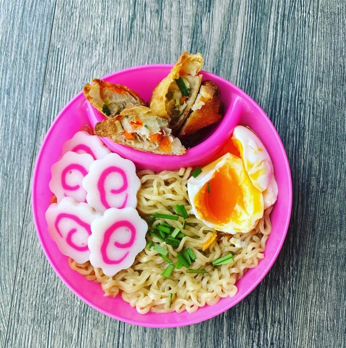 Bowl of ramen topped with a soft-boiled egg, sliced fish cake, and spring rolls on a textured surface