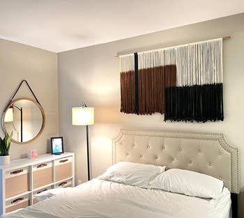 A reviewer’s photo of the black, brown, and white macrame wall hanging placed above their bed.