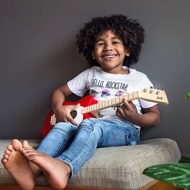 A child model playing a red and white Loog Mini acoustic guitar.