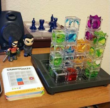 Puzzle game with clear towers placed on a table beside action figures and a Gravity Maze card deck. Chess pieces and a speaker are visible in the background.