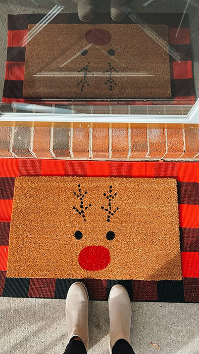 A reviewer standing in front of a Rudolph doormat, set on top of a red and black buffalo plaid rug