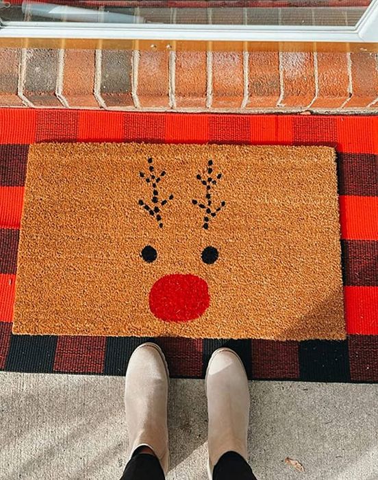 A reviewer standing in front of a Rudolph doormat, set on top of a red and black buffalo plaid rug