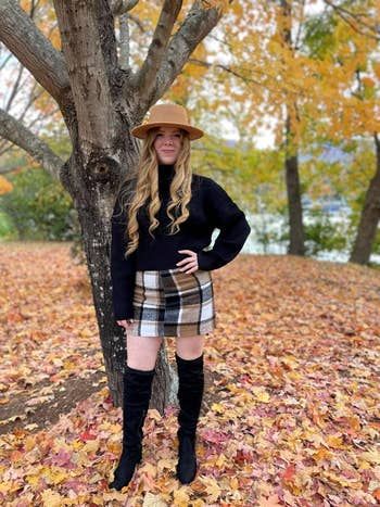 A reviewer stands outdoors amidst fall leaves, wearing a black sweater, plaid skirt, tan hat, and black over-the-knee boots.