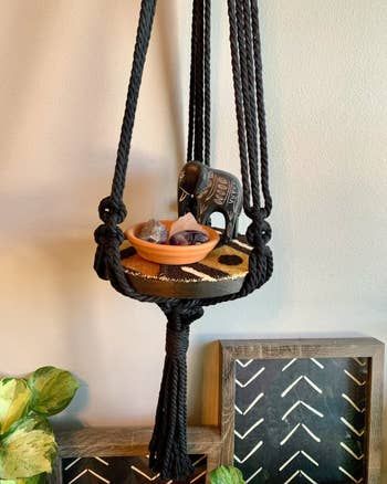 A macrame plant hanger displaying decorative items on a wall, with a framed artwork positioned below.