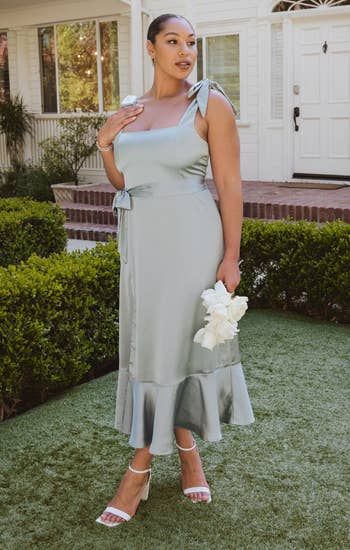 A stylish individual poses in a pastel midi dress with tie straps, holding white flowers, perfect for a sophisticated event or wedding