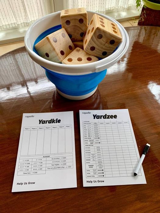 A set of Yardzee and Yardkle score sheets arranged on a wooden table, with a collapsible bucket containing large wooden dice. A black pen sits beside the sheets.