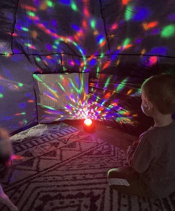 A child sitting on the floor, mesmerized by a light projector displaying colorful patterns in a dimly lit room