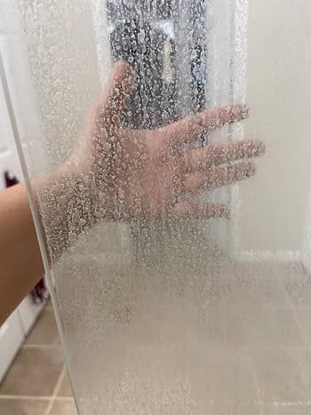 A reviewer’s hand partially hidden behind a glass shower door covered in cloudy spots