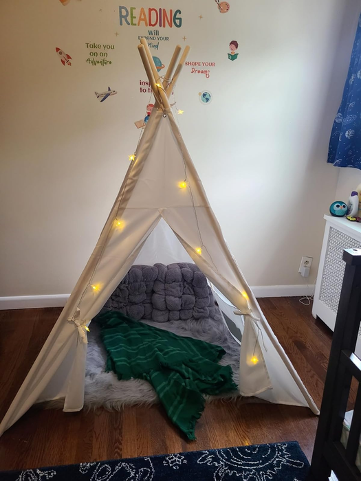 A children's play tent featuring string lights, plush cushions, and a green blanket, arranged in a cozy reading corner with motivational wall art