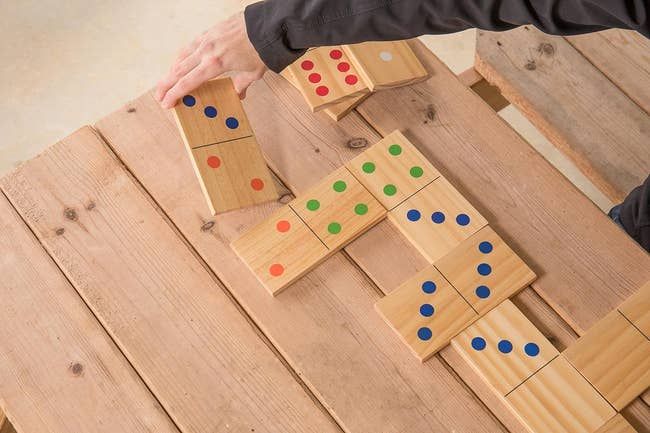 Someone arranges large wooden domino tiles, each adorned with colorful dots, on a wooden surface for an upcoming game.