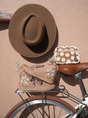 A hat, floral boots, and a matching floral bag rest on a bike rack with a plain background, featuring delicate white flowers.