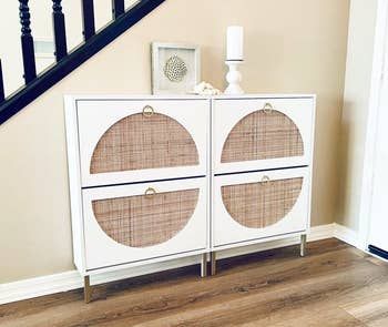 Two white, slim entryway storage units with rattan half-moon shaped designs and small gold handles that open up two compartments on each holder.