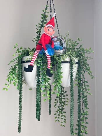 Elf doll perched on a hanging planter, holding a container labeled with a festive message