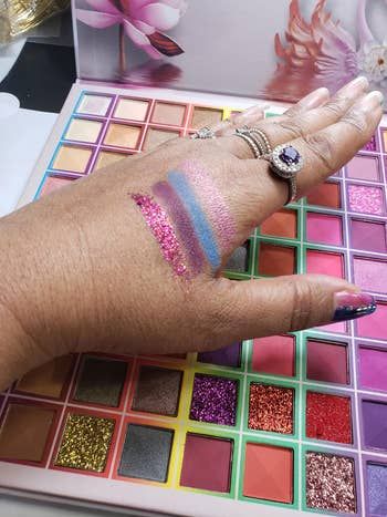 A reviewer’s hand showcasing four swatches of eyeshadow shades in front of an open palette filled with vibrant, multicolored options.