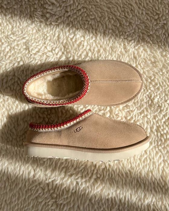 A pair of UGG slippers resting on a textured surface, showcasing cream-colored suede with a wooly interior and red accents along the edges.