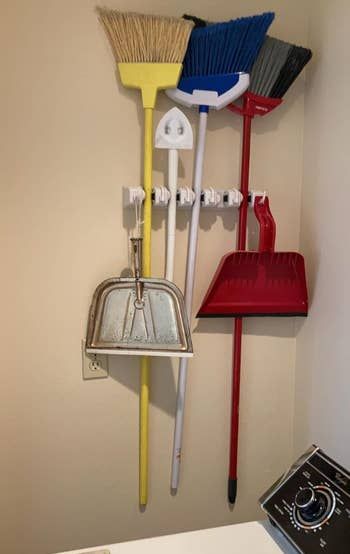 Various cleaning tools such as brooms, dustpans, and a mop are neatly hung on wall hooks inside a laundry room, with a washing machine corner visible.