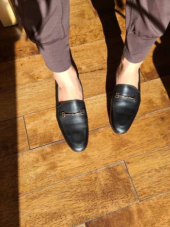Person in black loafers with metallic buckles, standing on a wooden surface