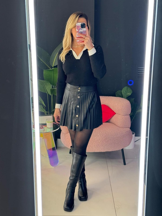 Person in a black sweater, pleated skirt, and tall boots posing in front of a mirror