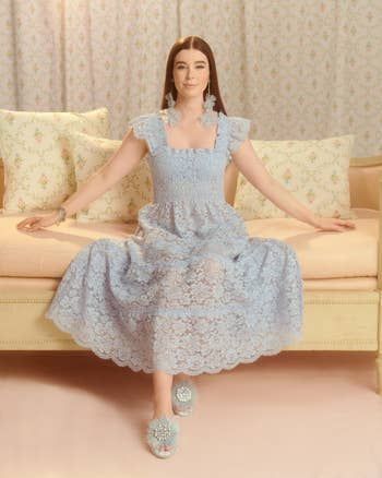 A person looking refined in an elegant lace dress, seated on a patterned sofa, accessorized with statement earrings and decorative sandals.