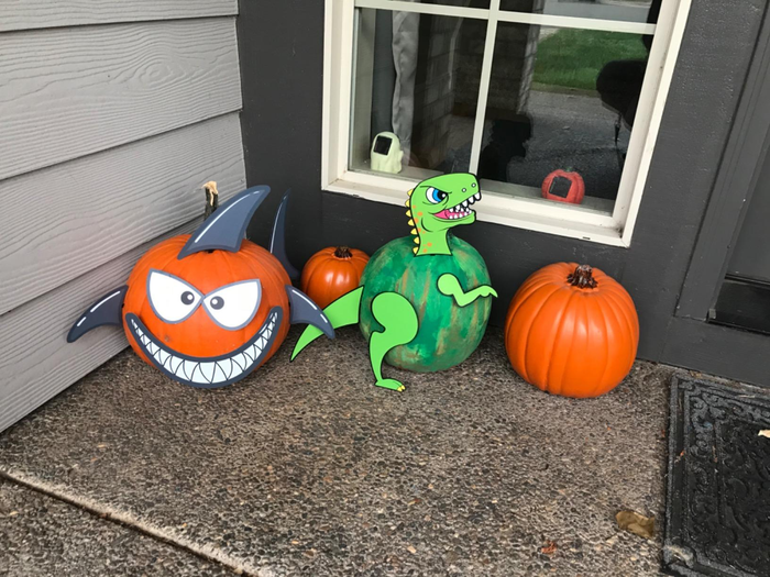 Three creatively adorned pumpkins placed by a front entrance: one featuring a shark design, another green pumpkin with a dinosaur silhouette, and a classic orange pumpkin