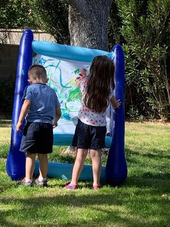 Two kids painting on a standing inflatable canvas in an open grassy area.