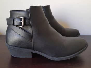 a reviewer's photo showcasing a pair of black ankle boots with a small block heel and a buckle design along the back heel.