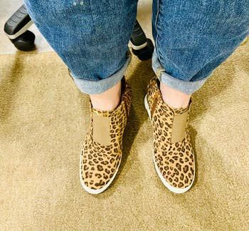 Leopard print slip-on shoes paired with rolled-up blue jeans, with feet resting near a chair.