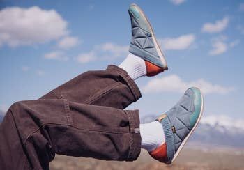 A person wearing casual shoes with white socks and dark pants, extending their legs toward the sky in an outdoor setting with mountains in the backdrop.