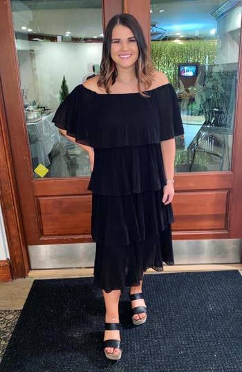 A reviewer wearing a tiered black off-the-shoulder dress paired with open-toe sandals, standing inside.