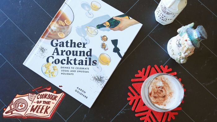 The 'Gather Around Cocktails' book placed on a table next to a glass of eggnog.