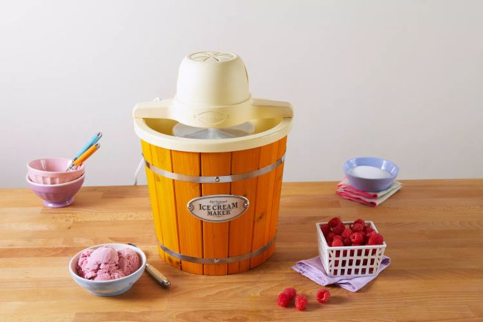 The Nostalgia 4-Quart Wood Bucket Ice Cream Maker displayed on a wooden counter next to a bowl of ice cream and fresh raspberries